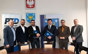 Na sali konferencyjnej stoi 6 osób. Osoby znajdujące się w środku trzymają teczki i gratulują sobie.